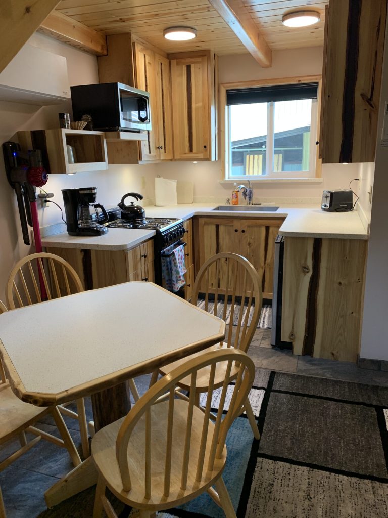 A modern kitchen featuring wooden cabinets, a gas stove, microwave, coffee maker, and a small dining table with chairs. Natural light fills the space through a window.