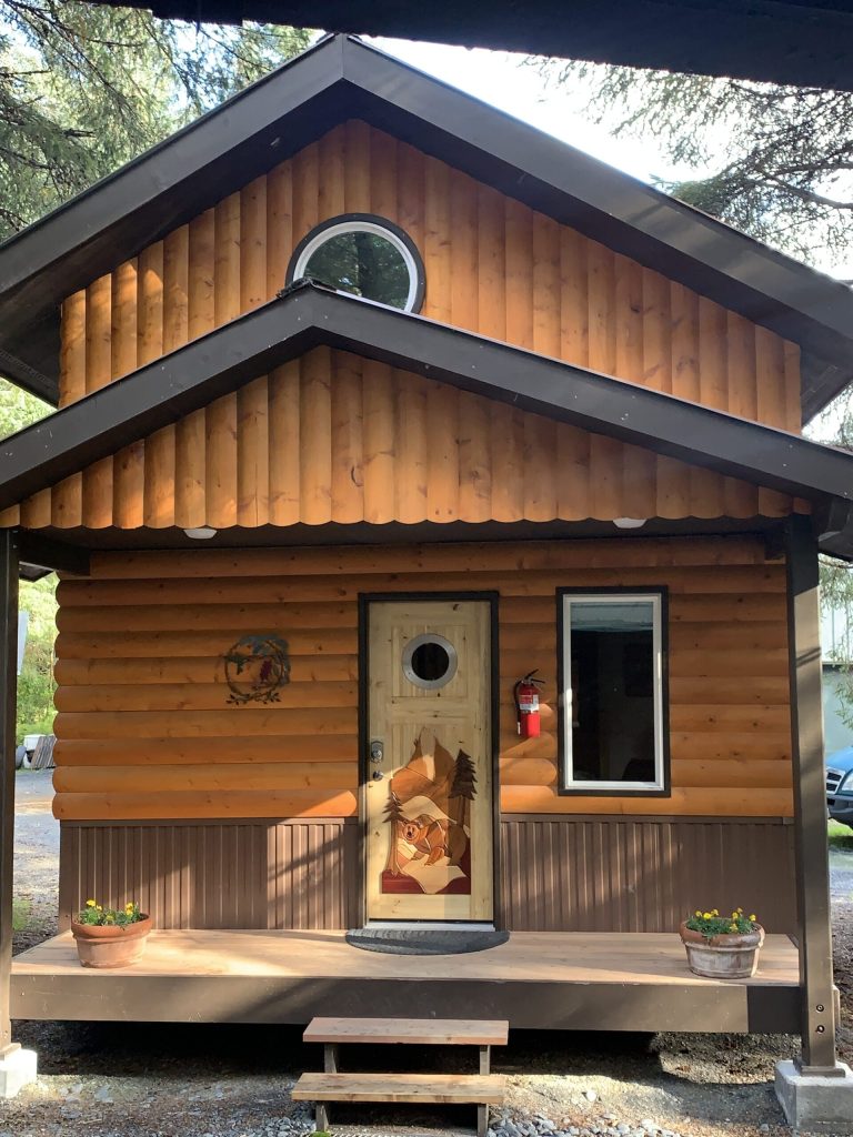 A charming wooden cabin with a sloped roof, featuring a circular window and a decorative front door. Potted flowers are placed on the porch, and there is a fire extinguisher near the entrance.