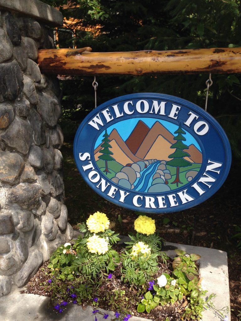 Welcome sign for Stoney Creek Inn featuring mountains, trees, and a river illustration, surrounded by flowers.