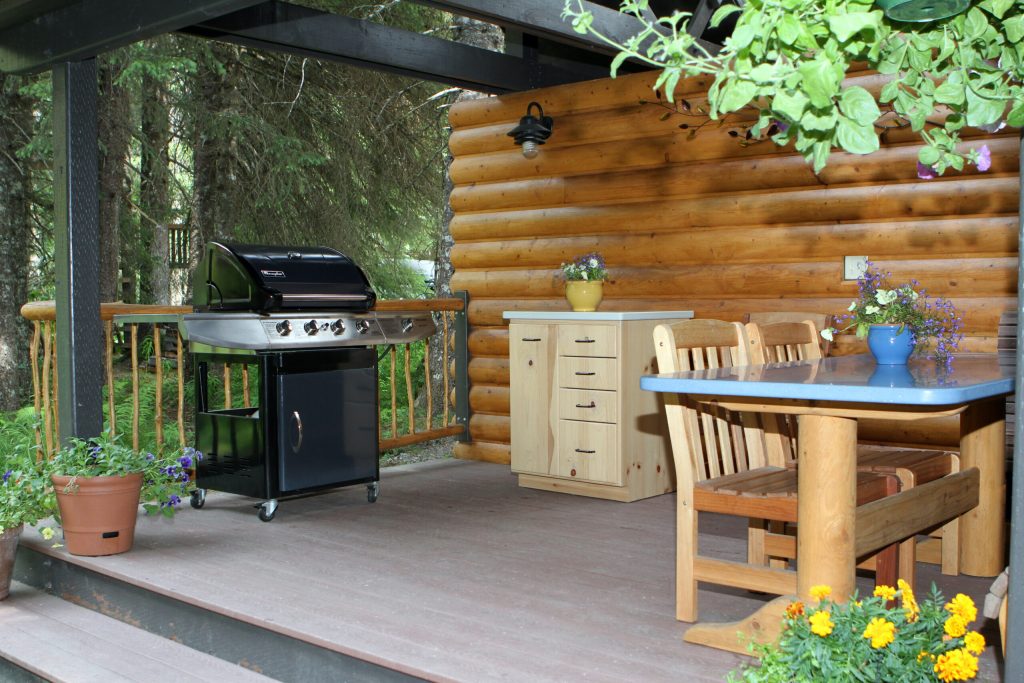 Outdoor patio area featuring a black gas grill, wooden dining table with chairs, a wooden cabinet, and potted plants under a log cabin structure.