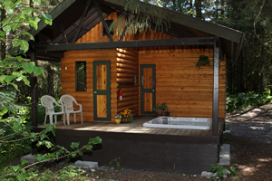 A cozy wooden cabin nestled in a forest, featuring a covered porch, two white chairs, and a small hot tub on the deck.