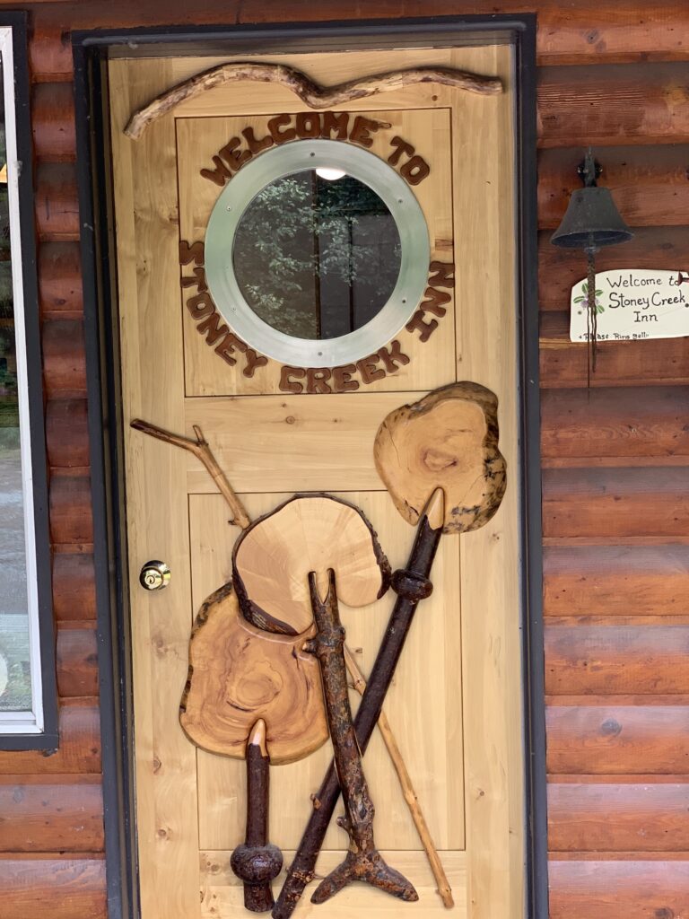 Wooden door with a circular window featuring the text 'Welcome to Stoney Creek Inn' carved above it, decorated with natural wood pieces and twigs.