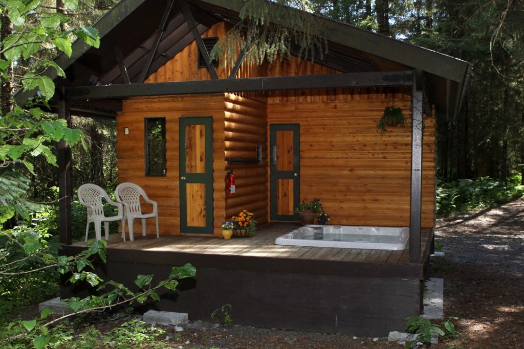 A wooden log cabin with a porch, featuring two white chairs, a small potted plant, and a hot tub, nestled in a forested area.