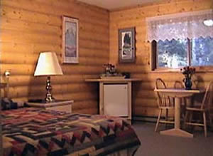 Cozy guest room with wooden log walls, a bed with a patterned quilt, a bedside lamp, a small table with chairs, and a window with lace curtains.