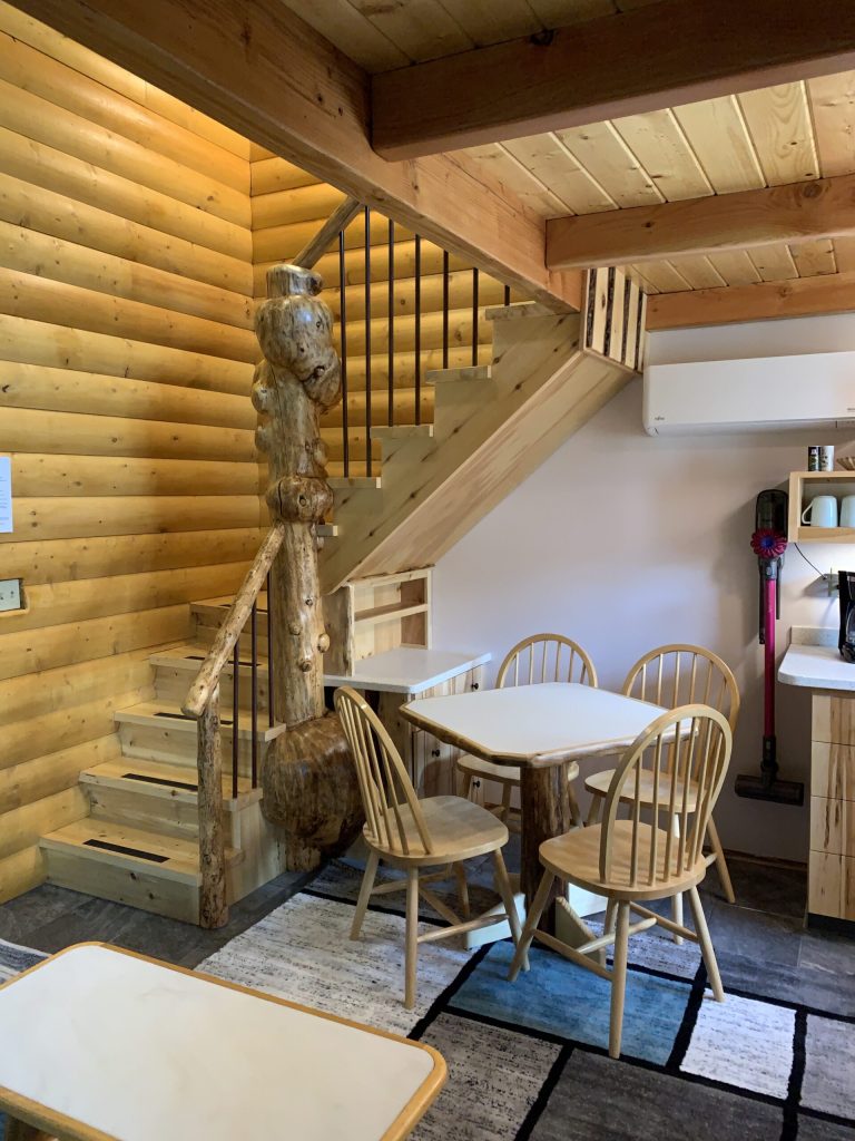 Cozy interior of a cabin featuring wooden walls, a staircase made of logs, and a dining area with round wooden chairs and tables.